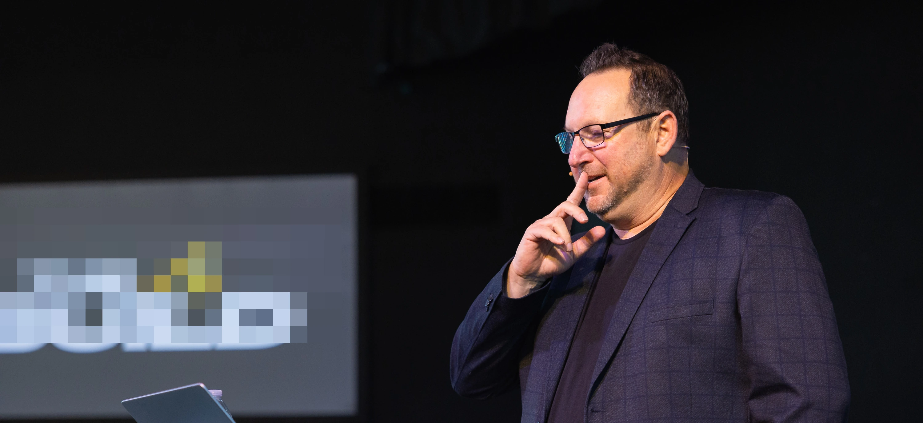 Michael Fineman on stage in a dark checked blazer, glasses on, a finger to his chin, mid-thought