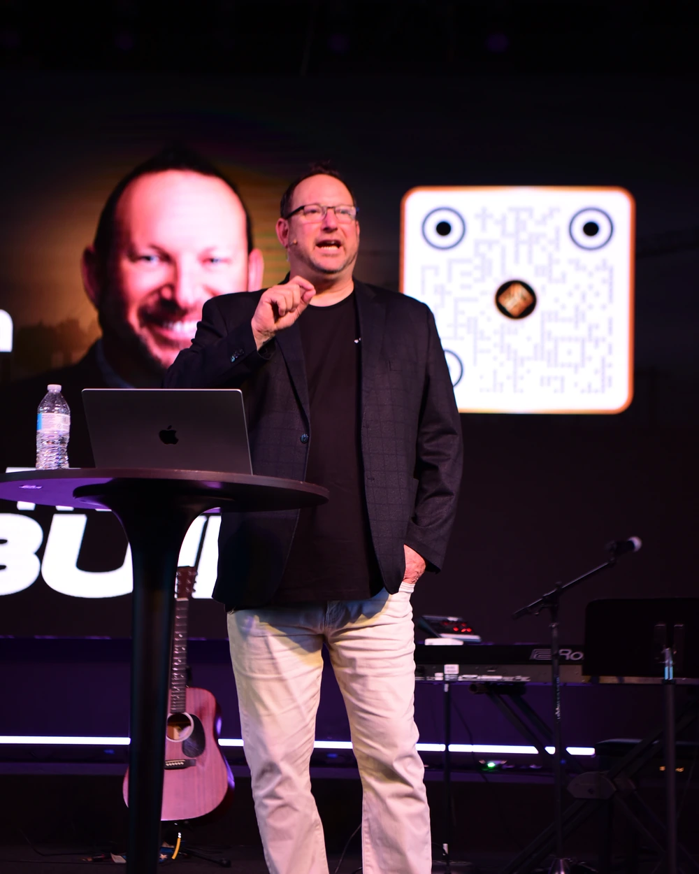 Michael Fineman on stage at The Build, mid-sentence, his face on the screen behind him