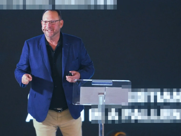 Michael Fineman smiling mid-keynote in a blue blazer, headset mic on, beside a clear lectern
