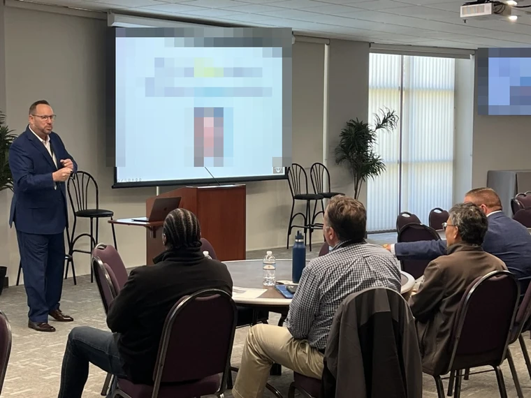 Michael Fineman presenting beside a projected slide to a small group seated at round tables